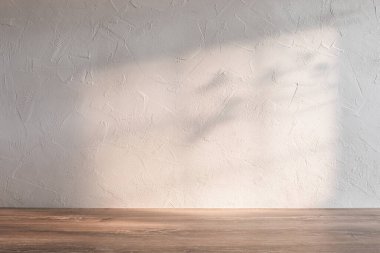 Sıcak bir güneş ışığı desenli bir duvarda ve ahşap bir masanın üstünde narin gölgeler oluşturarak sakin ve minimalist bir atmosferi vurguluyor..