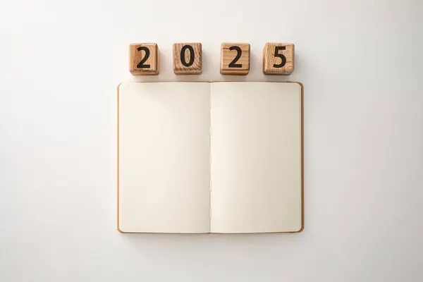 Boş defter masanın üzerinde duruyor, yanında 2025 numaralı tahta bloklar var, gelecek yıl için yeni başlangıçları, planları, hedefleri simgeliyor..