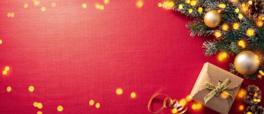 Çok güzel düzenlenmiş bir tatil ortamında altın bir fiyonkla süslenmiş parlak bir hediye kutusu, çam dalları, süsler ve zengin kırmızı bir zemin üzerinde ışıldayan ışıklarla çevrili..