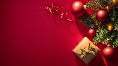 Güzel paketlenmiş bir Noel hediyesi, parlak süsler ve çam dallarıyla çevrili canlı kırmızı bir arka planda oturur, bayram neşesi ve kutlamaları çağrıştırır..