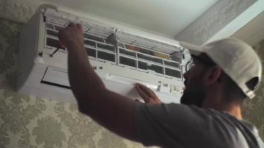 Odanın duvarındaki beyaz klima ünitesine tornavida kullanarak odaklanmış genç bir tamirci. Gri iş giysisi tamirinde uzman. Yüksek kalite 4k görüntü