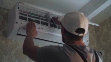 Odaklanmış genç tamirci odadaki duvardaki beyaz klima ünitesinde tozları fırçayla temizliyor. Gri iş kıyafetleri tamiri uzmanı. Yüksek kalite 4k görüntü