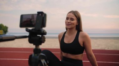 Spor blogcusu tripoddaki bir telefonla video dersi kaydediyor. Kumsalda, oyun alanında kamerayla konuşan bir kadın. Yüksek kalite 4k görüntü