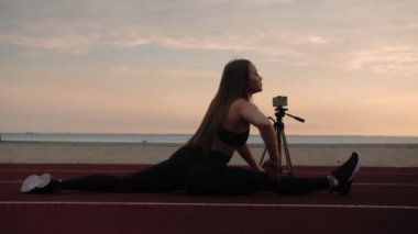 Fitness kız blogcusu sicimde oturuyor ve internette telefon kullanarak ders veriyor. Kumsalın önünde uzaktan çalışan bir kadın. Gün batımı. Yüksek kalite 4k görüntü