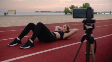 Spor sahasında karın kası egzersizi yapan bir kadın. Spor blogcusu tripoddaki bir telefonla video dersi kaydediyor. Açık hava fitness antrenmanı. Yüksek kalite 4k görüntü
