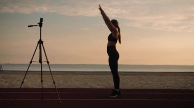 Spor giyimli genç bir kadın çevrimiçi fitness kursunda. Ters takla atıyor, gün doğumunda akrobatik hareketler yapıyor. Plajda spor öğreten bir kadın. Yüksek kalite 4k görüntü
