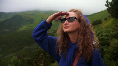 Dağlarda duran kadın mesafeye bakıyor. Kıvırcık saçlı kız manzaranın tadını çıkarıyor. Sırt çantalı mavi kapüşonlu kadın bir dağa tırmanıyor. Yüksek