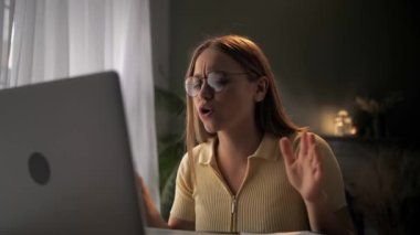 Genç bayan uzaktan kumandalı ofis çalışanı, dizüstü bilgisayarla masada oturuyor. Web kamerası kullanıyor, tartışıyor, patlıyor ve tartışıyor. Kızgın ve tatminsiz görünüyor. Kız çevrimiçi ağ kullanarak küfür ediyor. Yüksek