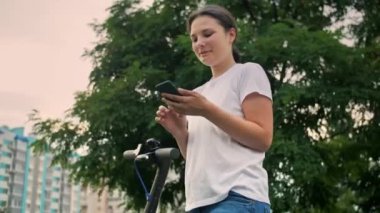 Şehirde e-scooter kullanan, akıllı telefon kullanan ve gülümseyen yüzü olan genç ve güzel bir kadın teslimat uygulaması kullanarak cep telefonu tutan bir kız. Yüksek kalite 4k görüntü