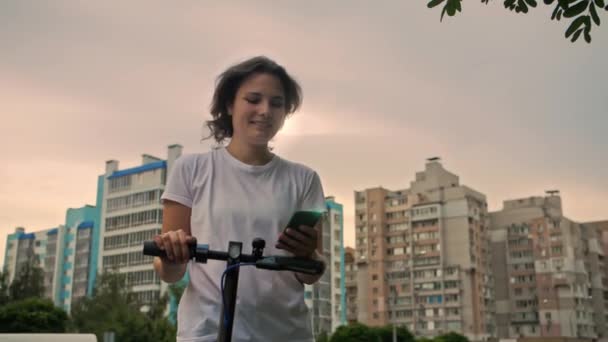 年轻美丽的女人站在城市里 拿着电动车 用智能手机和笑脸 迷人的女孩拿着手机 在汽车路上用投递应用程序 高质量的4K镜头 — 图库视频影像