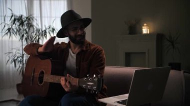 Erkek gitarist internetten bilgisayarla şarkı çalmayı öğreniyor. Adam akustik gitar çalıyor. Evde koltukta oturuyor. Genç sakallı adam enstrüman üzerinde akor çalışıyor. Yüksek kalite 4k görüntü
