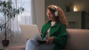 Kulaklık takan kadın evdeki koltukta oturup video görüşmesi yapıyor. Genç kıvırcık kız rahat koltukta dizüstü bilgisayar kullanıyor. Bilgisayarlı bir kadın internette öğrenim yapıyor. Yüksek kalite 4k