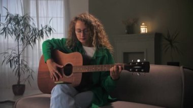 Evde koltukta oturan akustik gitar çalan bir kadın. Genç kıvırcık kız enstrüman üzerinde akor çalışıyor. Kadın gitarist akıllı telefonuyla şarkı çalıyor ve öğreniyor. Yüksek kalite 4k