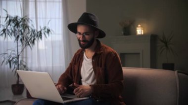 Adam evdeki koltukta oturup dizüstü bilgisayarla video görüşmesi yapıyor. Genç adam şapka takıyor, konuşuyor ve web kamerası yapıyor. Bilgisayarlı bir erkek internet üzerinden iletişim kuruyor. İş adamı toplantı konsepti yapıyor. Yüksek