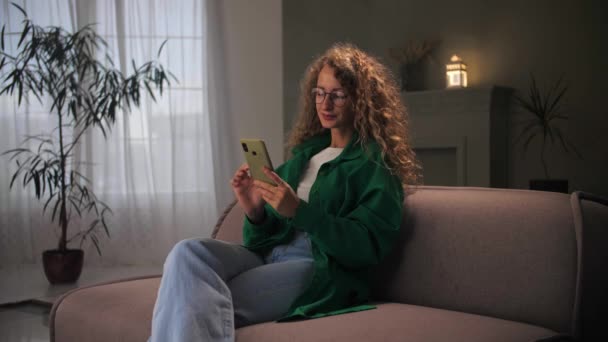女性はソファに座って電話を使用してインターネットを閲覧します スマートフォンの笑顔を見て若い巻き毛の女の子と家庭内でウェブサーフィン オンラインショッピングを行う女性は眼鏡を保持携帯電話を着用 — ストック動画
