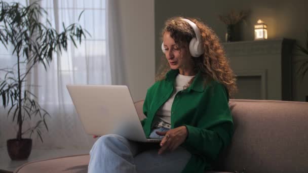 ヘッドフォンを着用した女性は 家庭内のソファに座ってビデオ通話を行います ラップトップを使用して快適なソファの上でリラックスした若い巻きガール オンライン学習を行うコンピュータを持つ女性 高品質4K — ストック動画