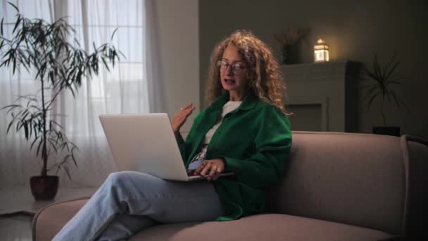 女性は自宅のインテリアでソファに座ってビデオ通話を行います ノートパソコンを使用して快適なソファの上でリラックスした若い巻き女の子 コンピュータがオンライン通信を行うと女性 — ストック動画