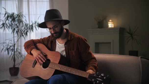 男人在家里的沙发上弹奏吉他 年轻的留胡子的男人在乐器上练习和弦 男性吉他手戴着帽子和眼镜 用记事本弹奏歌曲 — 图库视频影像