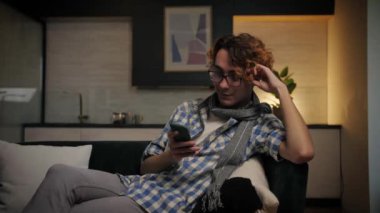 Kanepede uzanan genç adam telefon tarayıcı interneti kullanıyor.Rahat adam evde koltukta otururken tembelce yayın yapıyor. Gözlüklü, akıllı telefonlu ve kapalı alanda sörf yapan kıvırcık bir erkek. Yüksek