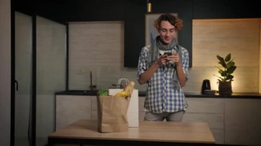 Delikanlı elinde poşetle mutfaktan eve geliyor. Alışveriş çantasındaki yiyecekleri kontrol ediyor. Süpermarket faturalarını cep telefonuyla kontrol ediyor. Yüksek kalite 4k görüntü