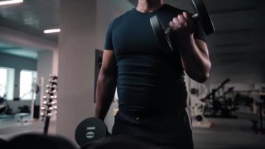 Spor salonunda halterli antreman yapan siyah bir adam. Vücut geliştirici kas geliştirici, biseps yapıyor, ellerinde ağır dambıllar tutuyor. Afrikalı Amerikalı Fitness elemanı ağırlık kaldırıyor. Yüksek kalite 4k