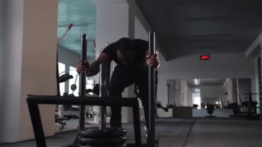 Siyah adam spor salonunda ağır bir yük vagonunu yavaş çekimde itiyor. Ağır bir ekipmanı ciddi yüzüyle iterek antrenman yapıyor. Yüksek kalite 4k görüntü