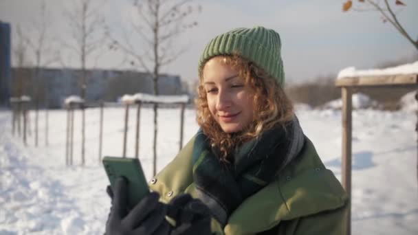 卷曲的女人在户外用手机浏览互联网 女孩拿着手机上网 脸上挂着微笑 冬天寒冷的背景 高质量的4K镜头 — 图库视频影像