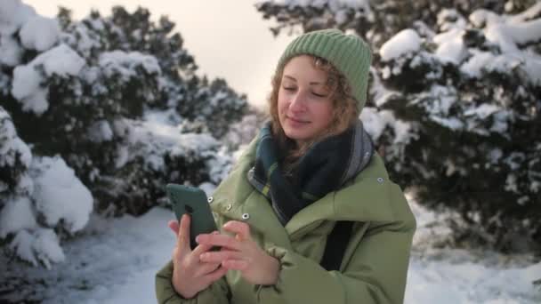 卷曲的女人在户外用手机浏览互联网 女孩拿着手机上网 脸上挂着微笑 冬天寒冷的背景 高质量的4K镜头 — 图库视频影像