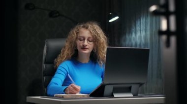 Kıvırcık kadın dizüstü bilgisayar ve not defteri kullanarak internette öğreniyor. Genç kız karanlık odada ciddi yüzleri olan notlar alıyor. İnternette İngilizce eğitimi alan bir kadın. Yüksek kalite 4k görüntü
