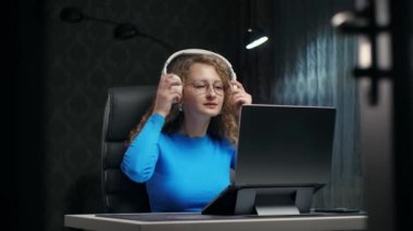 Genç Kıvırcık Sarışın Kadın kablosuz kulaklıklarla müzik dinlemeyi seviyor. Laptop 'ta çalışan ve karanlık ev içinde müzik dinleyen bir bayan. Yüksek kalite 4k görüntü