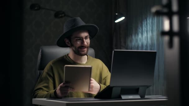 若いひげ男は暗い部屋の机に座ってノートパソコンを使用してオンラインで教える スマート男は帽子を着用する学生のためのオンライン教育を行うことを呼び出します家庭のリモート — ストック動画