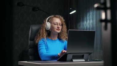 Genç Kıvırcık Sarışın Kadın kablosuz kulaklıklarla müzik dinlemeyi seviyor. Laptop 'ta çalışan ve karanlık ev içinde müzik dinleyen bir kadın müzik dinler ve daktilo çalar. Yüksek