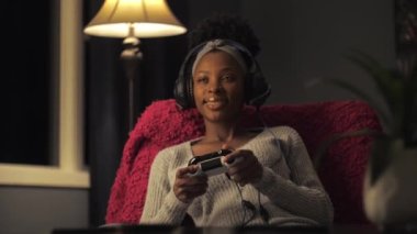 Afrikalı bir kadın karanlık oturma odasında oyun oynuyor. Genç bir kız evde online oyun oynarken kulaklık takıyor ve oyun platformu tutuyor. Siyah kadın mikrofonla konuşmaktan hoşlanıyor.