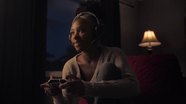アフリカの女性は暗いリビングルームに座ってジョイスティックを使用してゲームをプレイします 自宅でオンラインゲームをしている間 若い女の子はヘッドフォンを着用し ゲームパッドを保持します — ストック動画