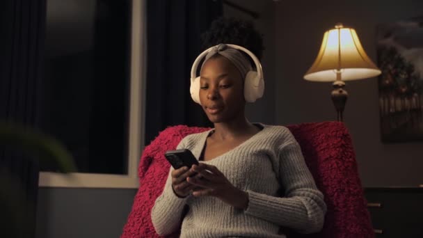 黒人女性はリビングルームの椅子に座ってヘッドフォンやスマートフォンを使って音楽を聴く 暖かい家庭のインテリアでスマートフォンを見てサーフィンインターネットを楽しんでいる若いアフロの女の子 — ストック動画