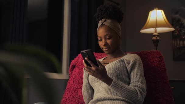 Czarna Kobieta Przeglądająca Internet Pomocą Telefonu Siedzi Ciemnym Wnętrzu Domu — Wideo stockowe