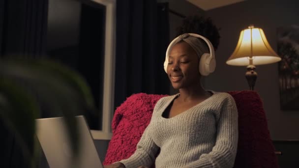 若いアメリカの女性は暗い家庭のインテリアで椅子に座ってノートパソコンの着用ヘッドフォンを使用してビデオ通話を行います ブラック女性は手を振って友人とのリモートオンライン会話を持っています — ストック動画