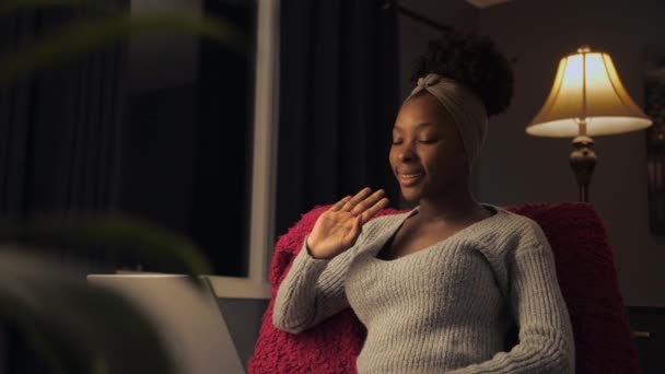 若いアメリカの女性は暗い家庭のインテリアで椅子に座ってラップトップを使用してビデオ通話を行います ブラック女性は手を振って友人とのリモートオンライン会話を持っています 高品質4K映像 — ストック動画