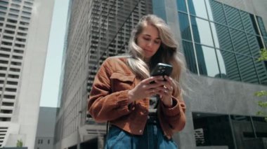 Sarışın bir kadın şehrin arka planında telefon taraması yapıyor. Kadın açık havada akıllı telefonlar yazan turuncu ceketli sörf sitesi takıyor. Cep telefonu bağımlısı insanlar. Yüksek kalite 4k görüntü