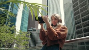 Genç sarışın kadın modern şehir binalarında fotoğraf çekiyor. Fotoğrafçı kadın fotoğraf makinesi kullanıyor. Fotoğraf eğitimi için kamera kullanıyor. Elinde aynasız fotoğraf makinesi olan bir kadın ekranlara bakıyor. Yüksek