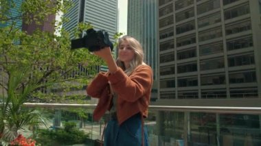 Genç bir kadın şehrin arka planında aynasız video kamera kullanarak video bloğu kaydediyor. Elinde kamera tutan sarışın kadın şehir merkezindeki izleyicileri etkiliyor. Çevrimiçi görüntü eğitimi. Yüksek kalite 4k
