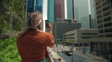Genç sarışın kadın fotoğraf çekiyor modern şehir binaları. Sarışın bayan telefonu kendi telefonu için kullanıyor. Akıllı telefonu olan kadın ekrandan bakıyor. Yüksek kalite 4k görüntü