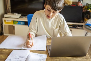 Çocuk evde dizüstü bilgisayar kullanıyor ve bir not defterine yazıyor, ödev yapıyor, eğitim sınavı konsepti