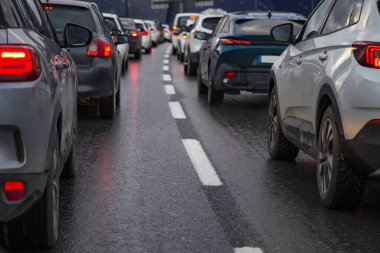 Akşamları iş çıkışı yoğun saatlerde otoyolda bir sıra araba ile trafik sıkışıklığı. Durmuş arabaların kırmızı fren lambaları gün batımında beyaz şeritli ıslak asfaltın arka planında.