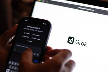 Teknisyen Grok 'u cep telefonuyla bilgisayar ekranında denerken Grok logosu gösteriyor. Elon Musk' un üretken, konuşkan, sinirsel bir ağı var.