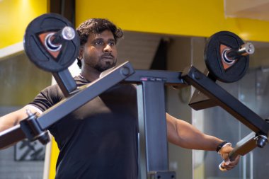 Omzunu güçlendirmek için omuz makinesinde beden eğitimi alan genç bir vücut geliştirici. Spor salonunda spor yaparken siyah tişört giyiyordu. Asya Hindistan spor salonu. Omuzlar için ayakta çalışma pozisyonu