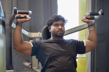 Omzunu güçlendirmek için omuz makinesinde beden eğitimi alan genç bir vücut geliştirici. Spor salonunda spor yaparken siyah tişört giyiyordu. Asya Hindistan spor salonu. Omuz egzersizi için oturma pozisyonu