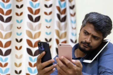 Sinirli ya da üzgün Asyalı Hintli adam birden fazla dijital cihaz kullanıyor.