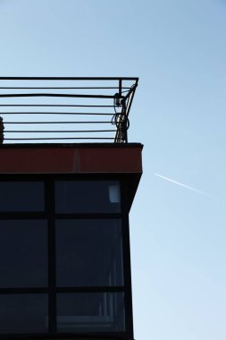 Part of a building with large glass windows and a blue sky, the path of the plane visible as a line in the sky