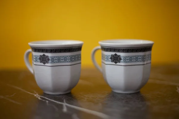 Two white coffee mugs with gray and blue details, yellow background and gray marble on which the mugs stand